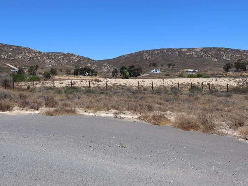 A view of a vacant residential plot featuring a wire mesh fence and access to an asphalt road, set against a natural landscape.