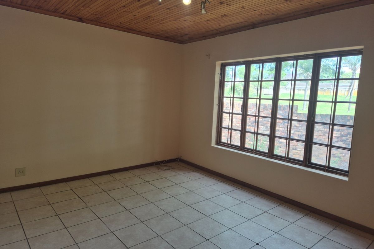An empty living room featuring ceramic floor tiles, a wooden tongue and groove ceiling, and a large window with security bars.