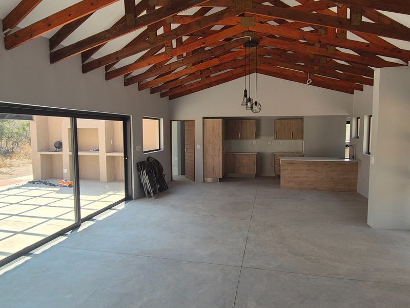 Spacious open-plan living area featuring exposed timber roof trusses, polished concrete floors, and a modern kitchen with an island, opening to an outdoor patio.