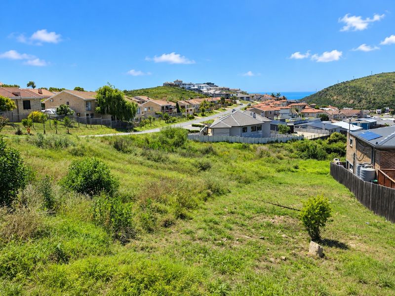 A view of a sloped residential vacant land plot overlooking a suburban neighborhood with distant coastal scenery.