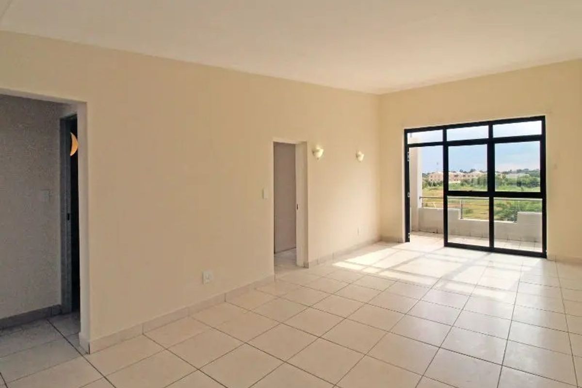 An empty, open-plan living area featuring neutral tiled flooring, wall-mounted lighting, and a large sliding glass door providing access to an outdoor balcony.