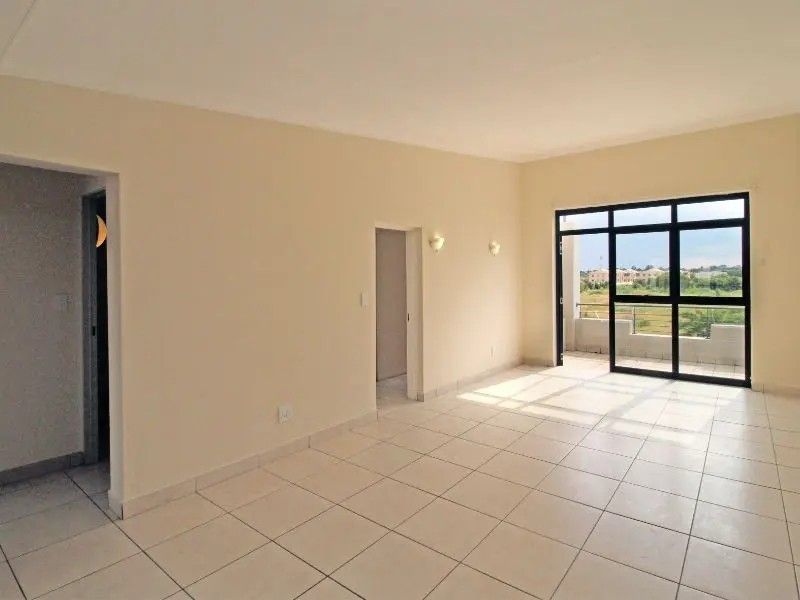 An empty, open-plan living area featuring neutral tiled flooring, wall-mounted lighting, and a large sliding glass door providing access to an outdoor balcony.