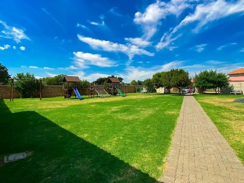 A well-maintained communal garden area featuring a paved walkway, expansive lawn, and established playground equipment enclosed by a perimeter brick wall.