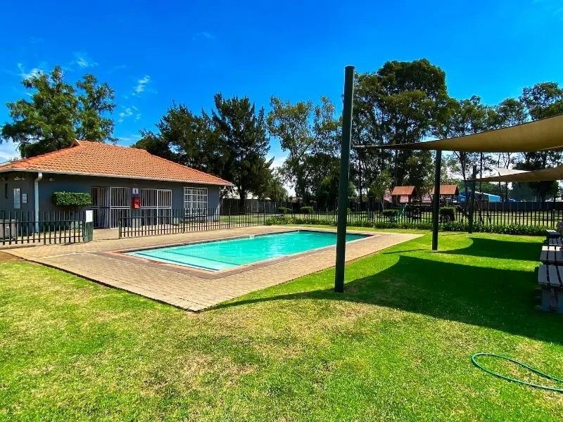 A well-maintained communal pool area featuring a paved deck, shade sail structure, and adjacent clubhouse building within a landscaped garden setting.