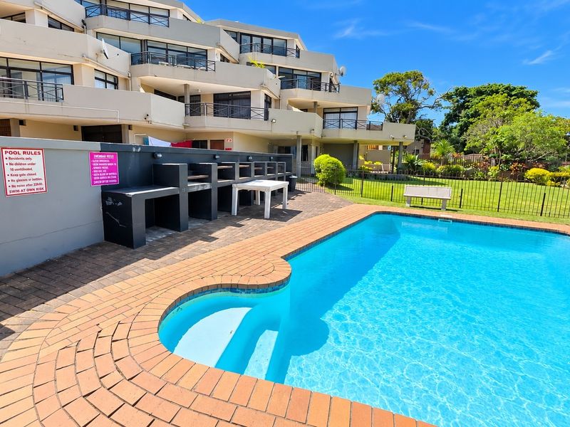 Communal swimming pool area featuring a paved deck, built-in braai facilities, and metal perimeter fencing, set within a well-maintained apartment complex garden.