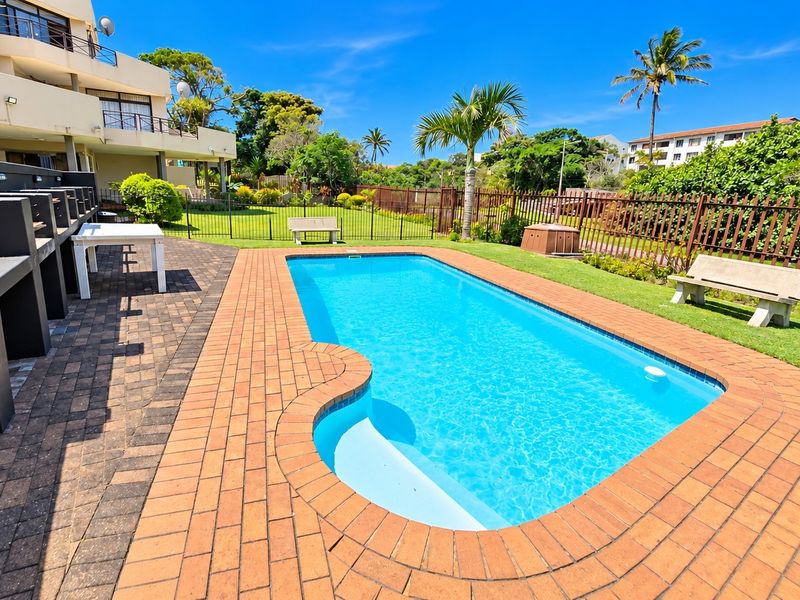 Outdoor pool area featuring a swimming pool, brick paved surround, built-in braai facility, and landscaped lawn within a fenced complex setting.