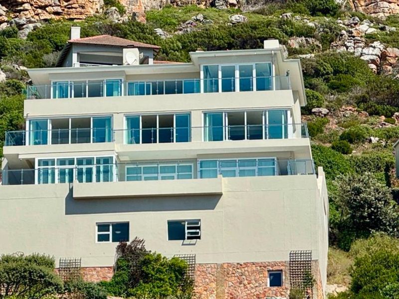 A multi-level residential property featuring extensive glass balustrades, sliding doors, and stone cladding, set against a natural mountain backdrop.