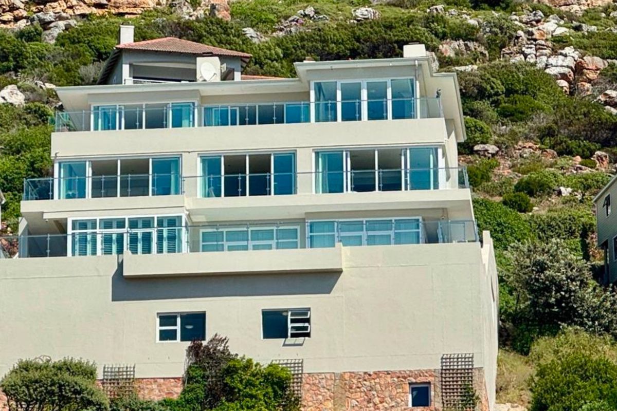 A multi-level residential property featuring extensive glass balustrades, sliding doors, and stone cladding, set against a natural mountain backdrop.