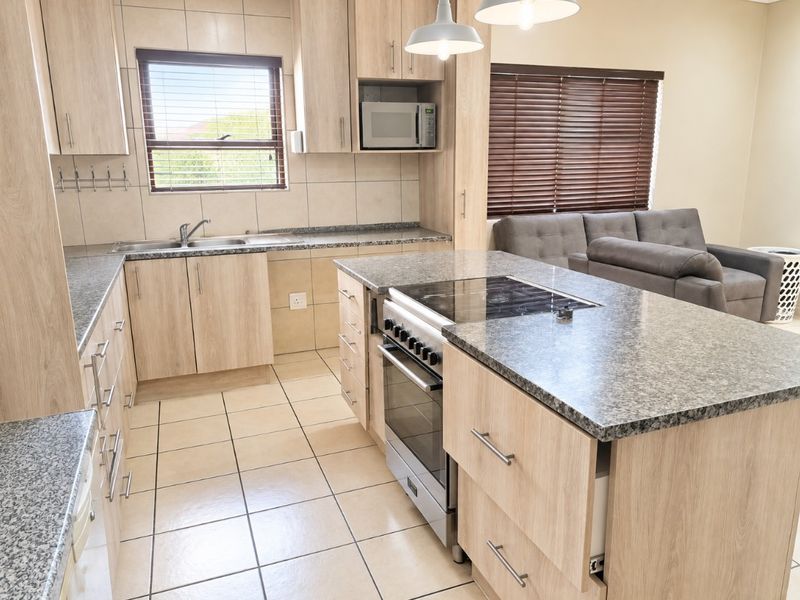 Modern open-plan kitchen featuring light wood cabinetry, granite countertops, a built-in oven and hob, and tiled flooring, seamlessly flowing into the living area.
