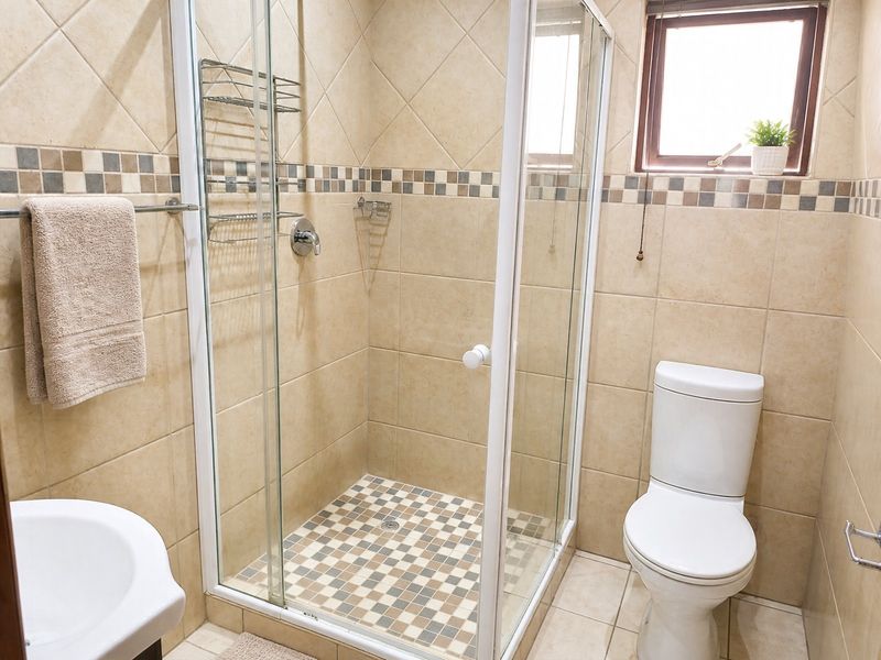 A functional bathroom featuring a glass-enclosed shower, toilet, and neutral-toned wall and floor tiling with a decorative mosaic border.