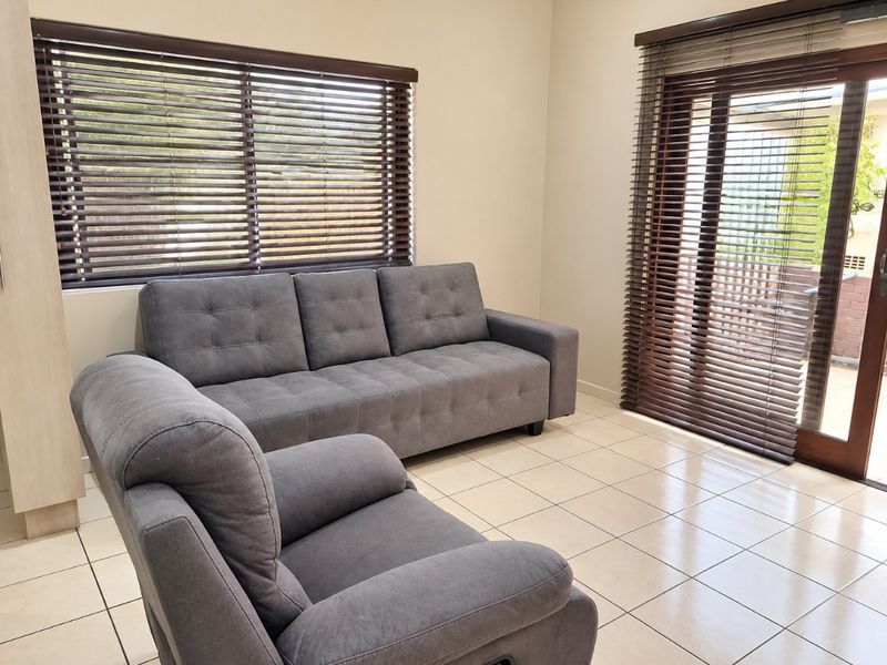 A bright living room featuring neutral tiled flooring, large windows with venetian blinds, and a sliding glass door providing access to the outdoor area.