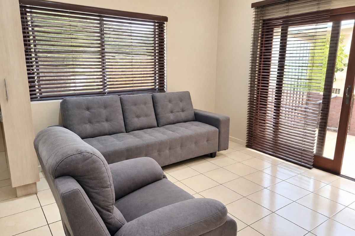 A bright living room featuring neutral tiled flooring, large windows with venetian blinds, and a sliding glass door providing access to the outdoor area.