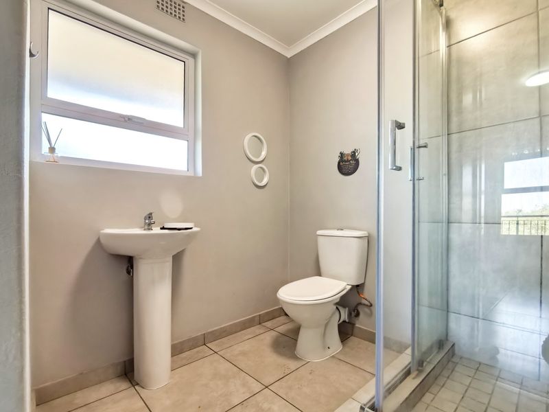 A clean and functional bathroom featuring a pedestal basin, a standard toilet, and a glass-enclosed shower with neutral floor and wall tiling.