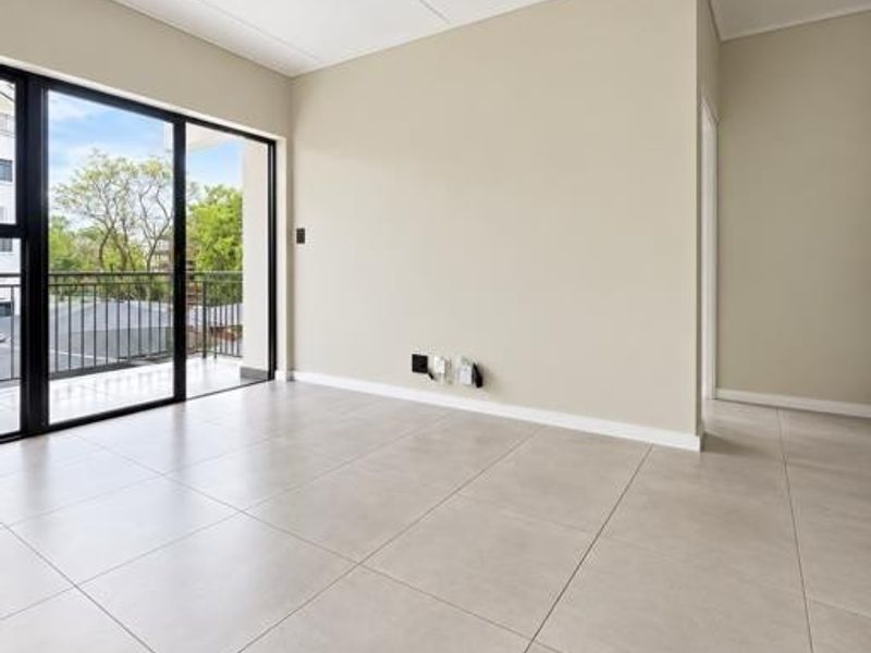 A bright, empty living area featuring large format floor tiles, modern track lighting, and a sliding glass door leading to a private balcony.