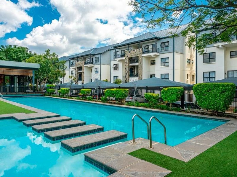 A well-maintained communal swimming pool area featuring stone coping, artificial turf, and views of the modern apartment complex with covered parking.
