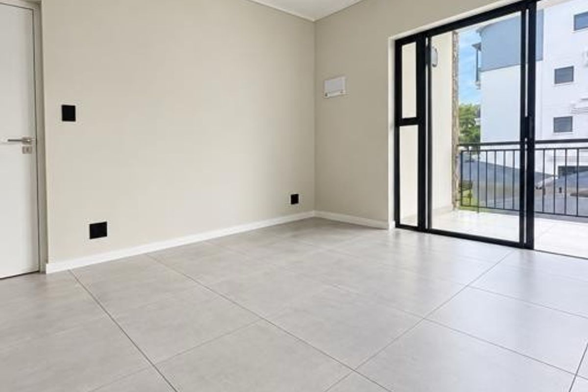 A bright, empty living room featuring large format floor tiles, modern track lighting, and a sliding glass door leading to a balcony.