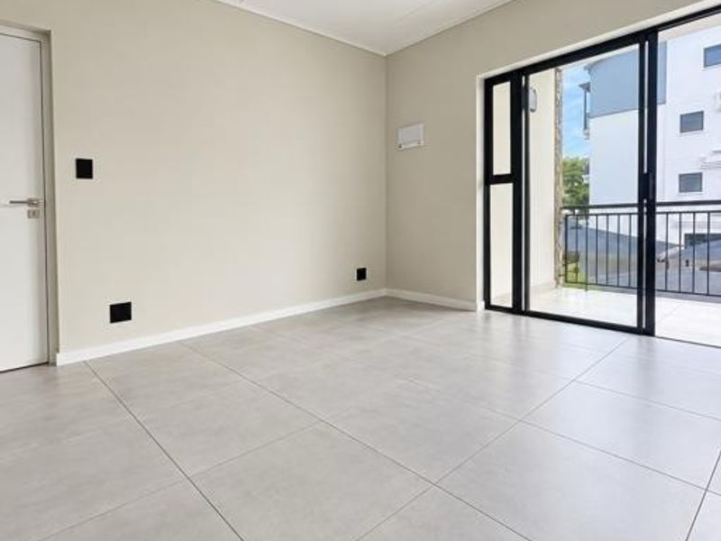 A bright, empty living room featuring large format floor tiles, modern track lighting, and a sliding glass door leading to a balcony.