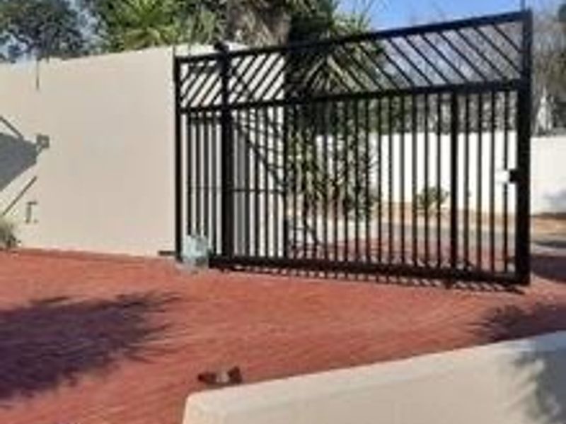 A secure front entrance featuring a black metal sliding gate, brick paved driveway, and perimeter boundary wall.