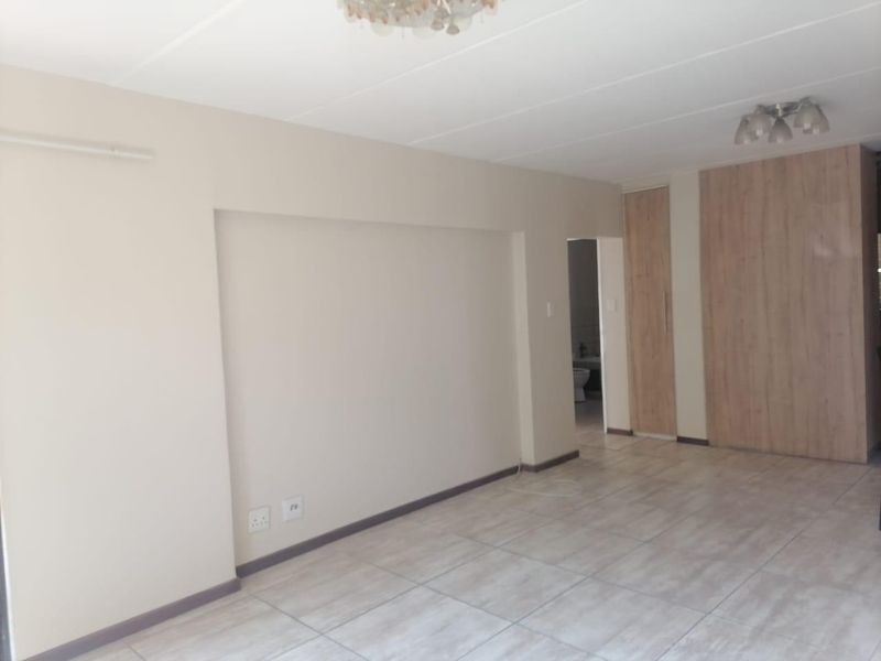 A spacious, empty bedroom featuring neutral wall tones, tiled flooring, and built-in wooden cupboards, offering a clean and versatile living space.