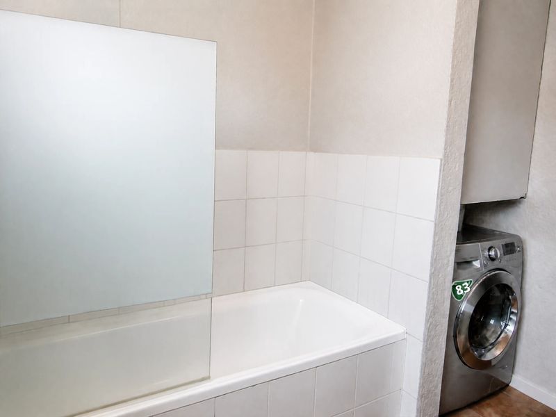 A functional bathroom featuring a built-in bathtub with a glass shower screen, tiled splashback, and a dedicated laundry area with built-in cabinetry.