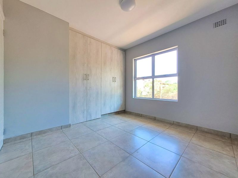 A bright, empty bedroom featuring neutral tiled flooring, built-in cupboards with a light wood finish, and a large window providing natural light.