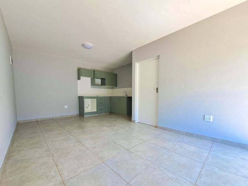An open-plan living area featuring a compact kitchen with sage green cabinetry, subway tile backsplash, and neutral tiled flooring throughout.