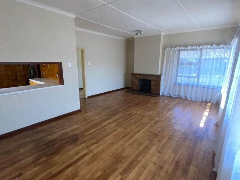 Spacious living room featuring original hardwood flooring, a classic brick fireplace, and a convenient serving hatch connecting to the kitchen area.