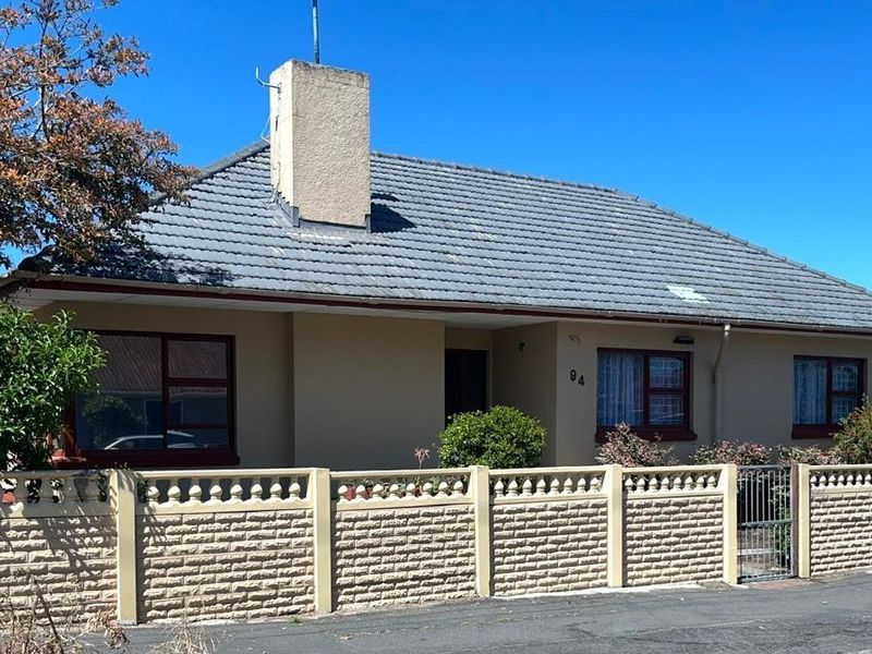 Front elevation of a single-story house featuring a tiled roof, chimney, and a secure precast concrete boundary wall with a pedestrian gate.
