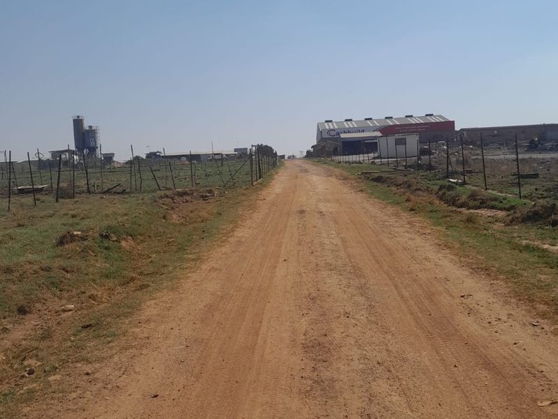 A view of a dirt access road leading towards an industrial farm property with fencing, a silo, and a large warehouse structure.