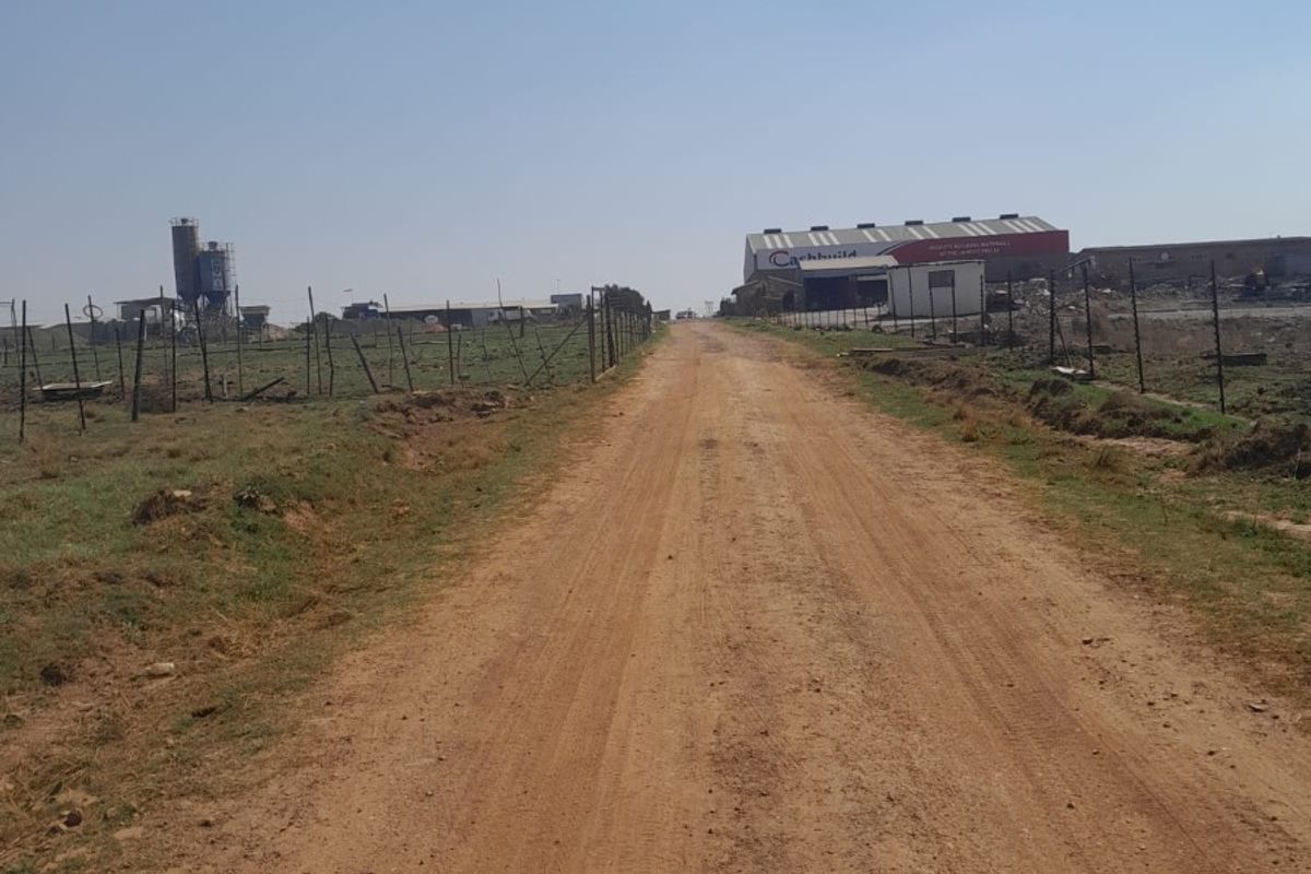 A view of a dirt access road leading towards an industrial farm property with fencing, a silo, and a large warehouse structure.