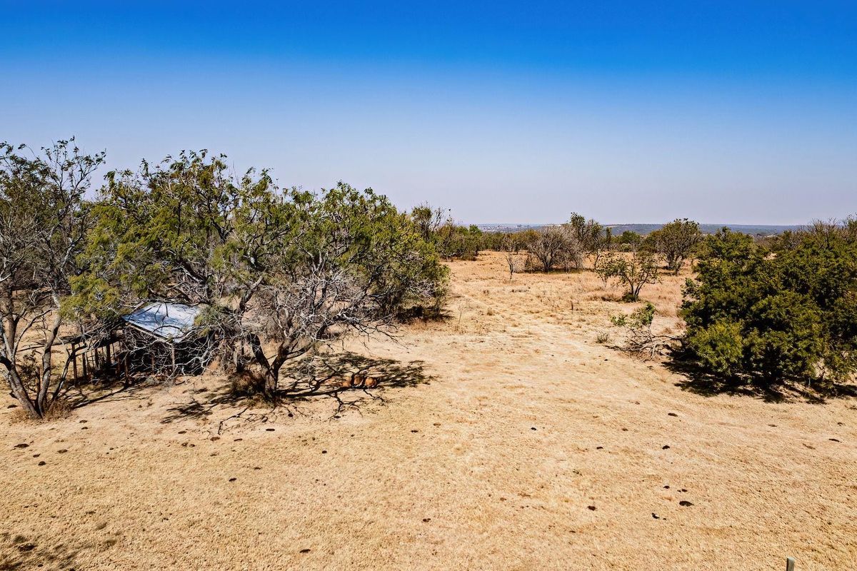 Aerial view of a large, dry farm landscape featuring mature trees and a small, weathered outbuilding with a corrugated metal roof.