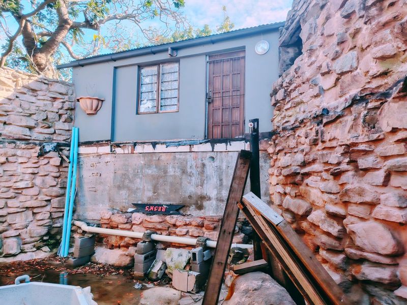 Rear exterior view showing a stone retaining wall, exposed foundation, and a small structure with a security gate and window.