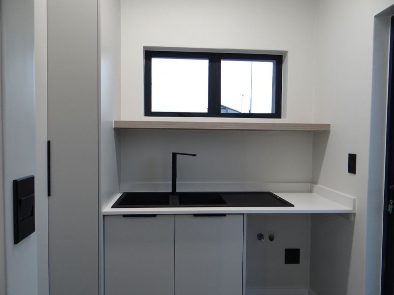 A modern, minimalist laundry room featuring sleek white cabinetry, a black sink with matching tapware, and a stone countertop with plumbing for appliances.