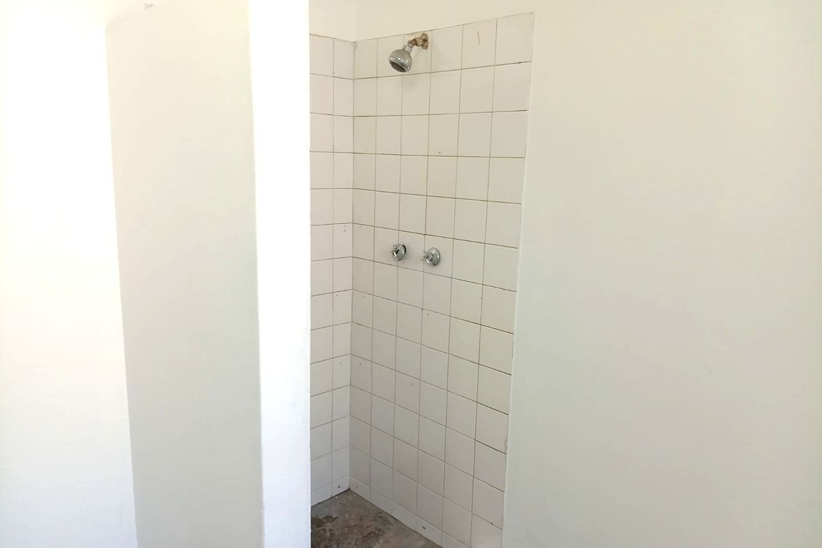 A basic shower enclosure featuring white wall tiles and exposed plumbing fixtures, currently requiring floor finishing and general maintenance.