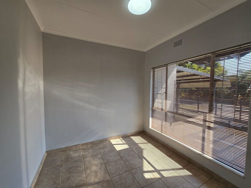 A bright, empty bedroom featuring neutral grey walls, tiled flooring, and large windows fitted with venetian blinds.