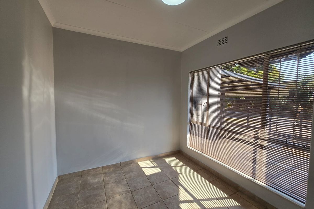 A bright, empty bedroom featuring neutral grey walls, tiled flooring, and large windows fitted with venetian blinds.