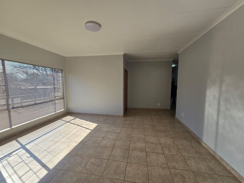 A bright, empty living room featuring neutral grey walls, tiled flooring, and large windows fitted with venetian blinds.