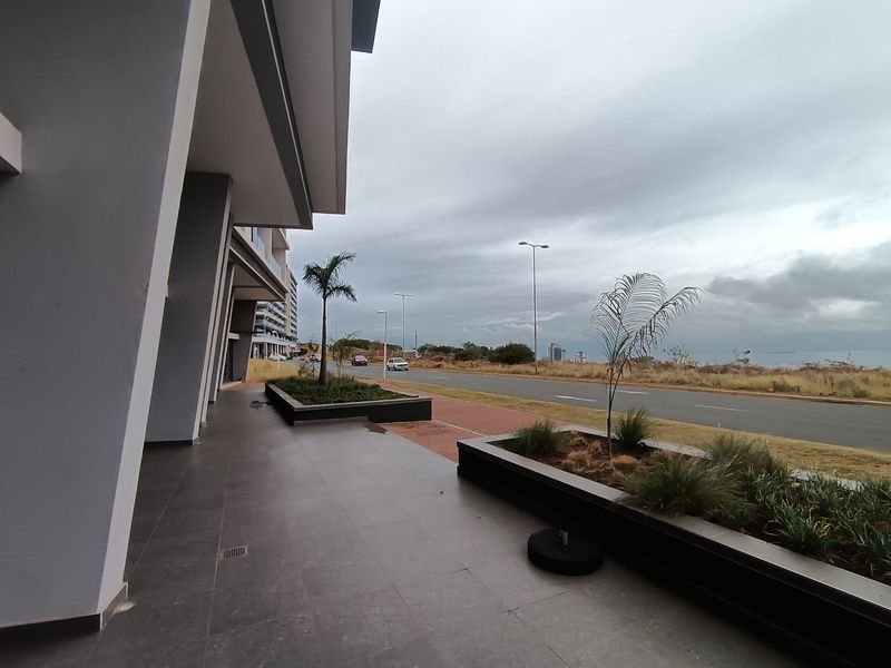 A modern commercial exterior walkway featuring tiled flooring, raised garden beds, and structural pillars overlooking a coastal road.