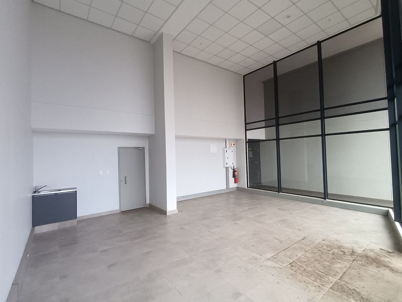 Spacious open-plan retail unit featuring high ceilings, large glass windows for natural light, tiled flooring, and a compact kitchenette area.