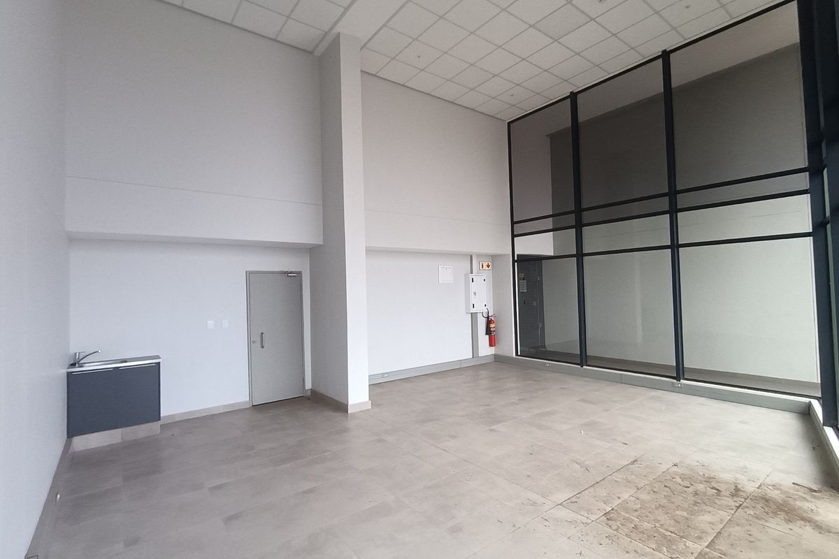 Spacious open-plan retail unit featuring high ceilings, large glass windows for natural light, tiled flooring, and a compact kitchenette area.