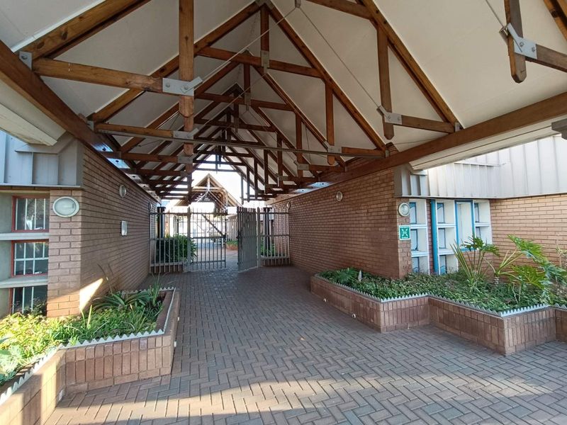 A covered entrance walkway featuring exposed timber roof trusses, brick masonry walls, and integrated brick planters leading to a secure gated access point.