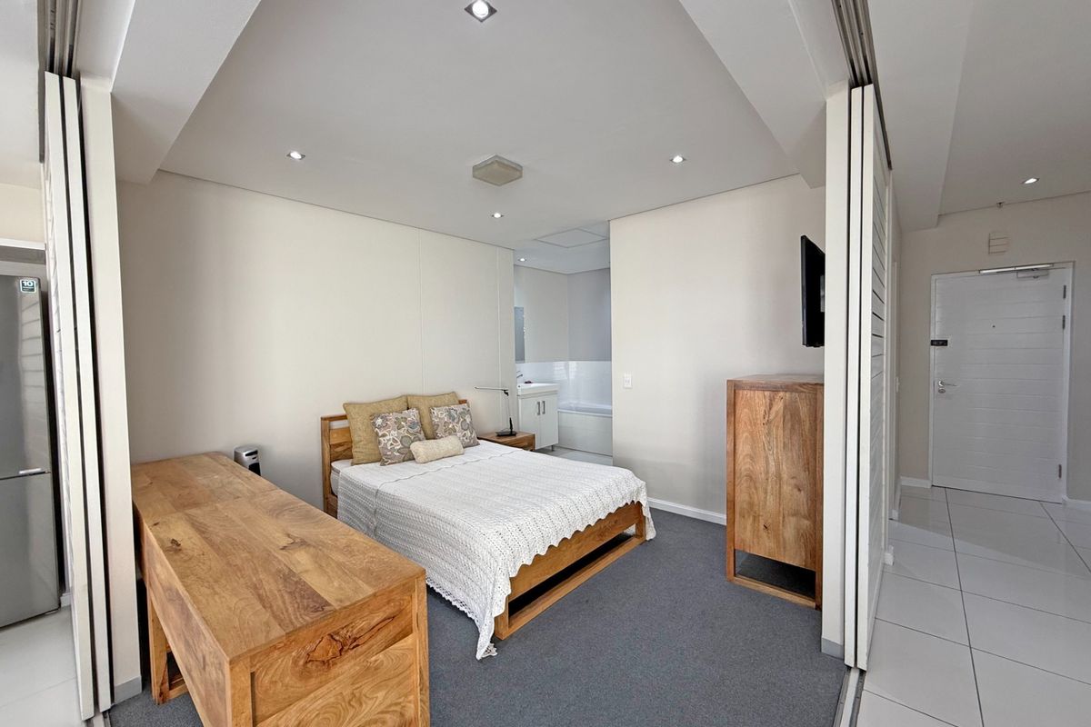 A well-lit bedroom featuring a neutral color palette, carpeted flooring, recessed lighting, and convenient access to an en-suite bathroom through sliding partition doors.