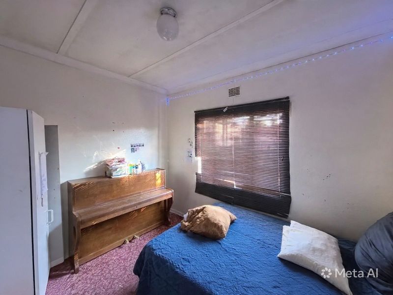 A bedroom featuring a window with venetian blinds, ceiling lighting, and ventilation, currently showing signs of wear on the walls and flooring.