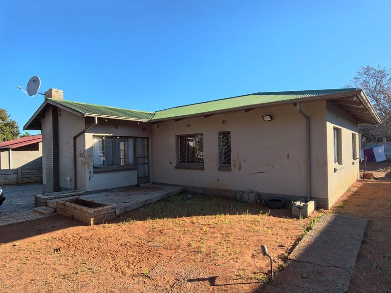 Rear view of a single-story house featuring a corrugated metal roof, brick chimney, and a small concrete patio area with surrounding garden space.