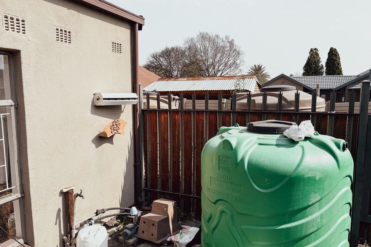 Rear exterior area featuring a large water storage tank, water pump, and external tap against a plastered wall with metal perimeter fencing.