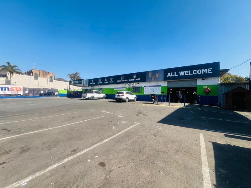 Front exterior view of a commercial retail property featuring a large asphalt parking area and a roller shutter entrance.