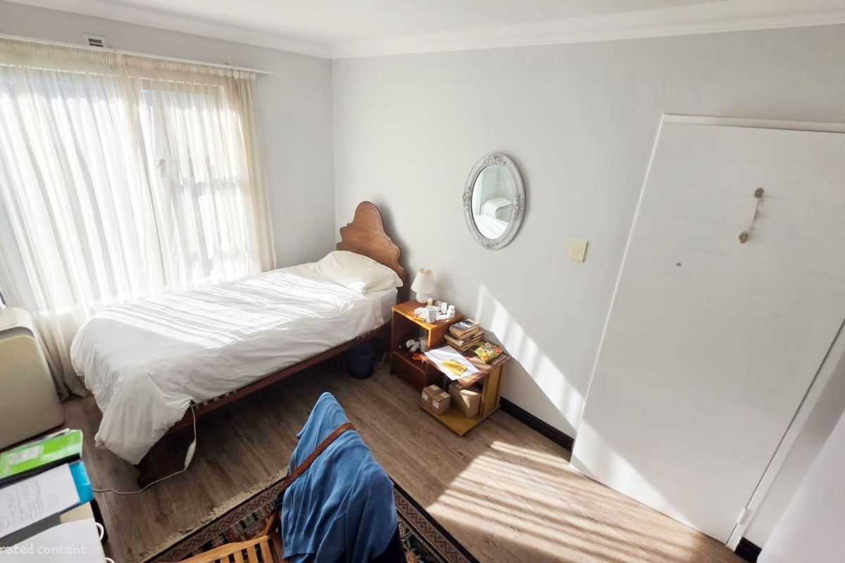 A functional bedroom featuring laminate flooring, a built-in wardrobe, and natural light through a large window with sheer curtains.