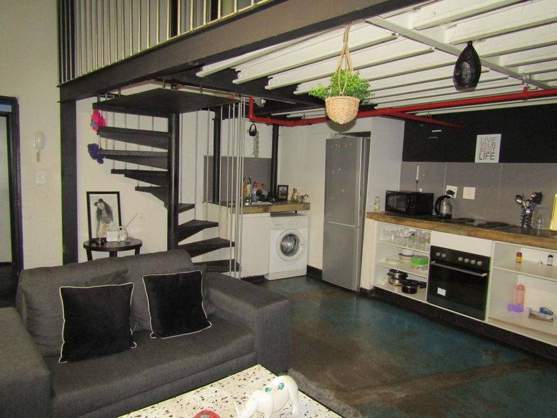 Open-plan living area featuring an industrial aesthetic with a spiral staircase, mezzanine level, exposed steel beams, and a compact kitchen setup.