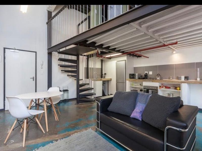 Industrial-style open plan living area featuring a mezzanine level, spiral staircase, exposed steel beams, and a functional kitchen with built-in cabinetry.