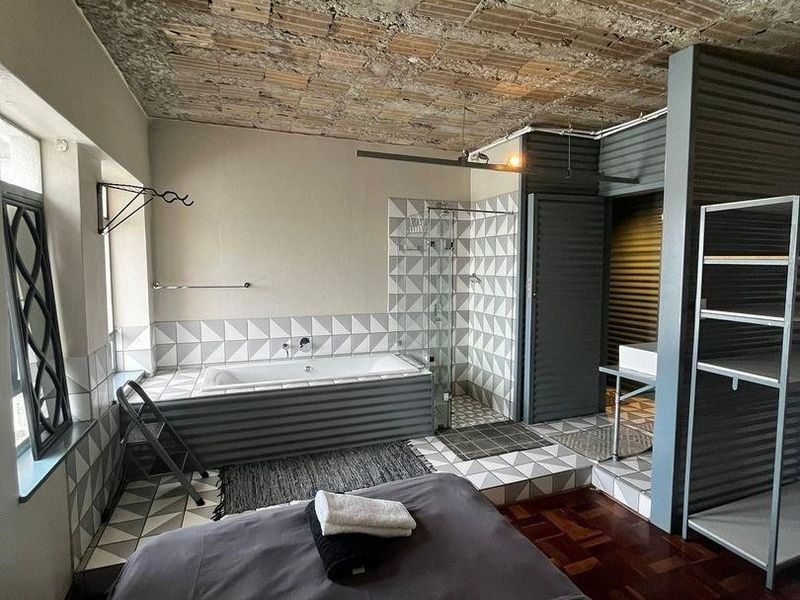 Unique bedroom featuring an open-plan ensuite with a built-in bathtub, glass shower, exposed brick ceiling, and industrial-style corrugated metal accents.